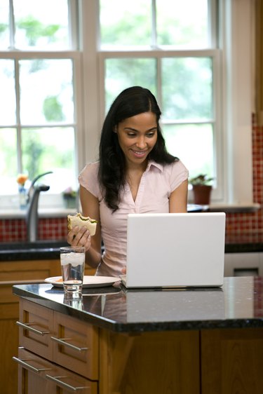 Woman on laptop in kitchen, eating snack